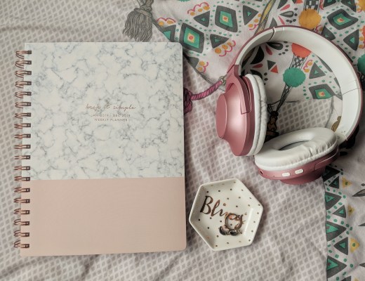An open journal with a marbled and pink cover titled "Weekly Planner: March Edition" lies on a patterned bedsheet next to folded pink and white headphones. A hexagonal dish with the word "Hello" and a ring inside it rests nearby.