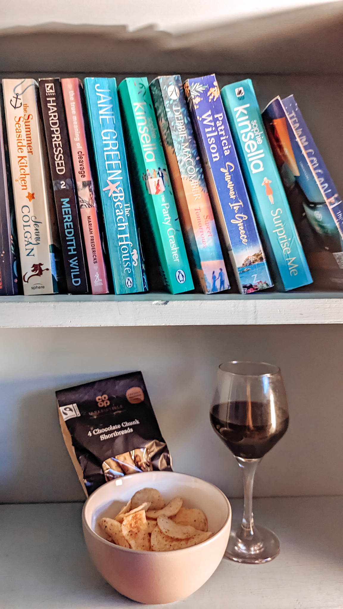 A glass of wine, a small bowl of crisps and a packet of cookies on a bookshelf filled with books
