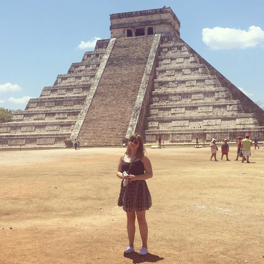 Formidable Joy - UK Fashion, Beauty & Lifestyle Blog | Travel | My Mexican Adventure | Day One - Chichen Itza & Merida Louise standing in front of Chichen Itza in Mexico! She is wearing a dark blue vest top with a knee-length skirt, which is patterned with stripes in different shades of blue. Behind her, the iconic stone pyramid of Chichen Itza rises above her.