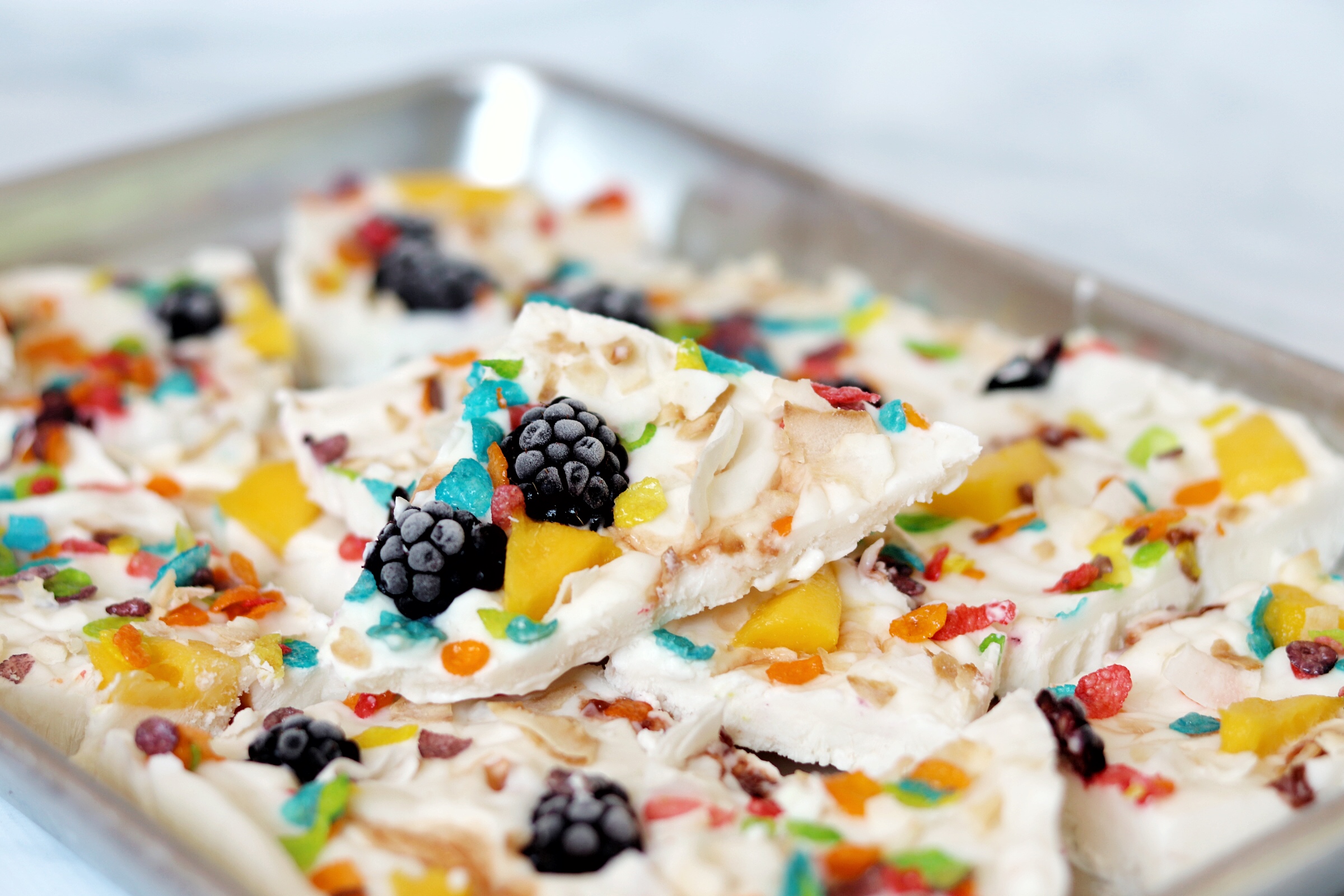 Frozen yogurt bark topped with fruit and Fruity Pebbles cut into squares in a baking pan