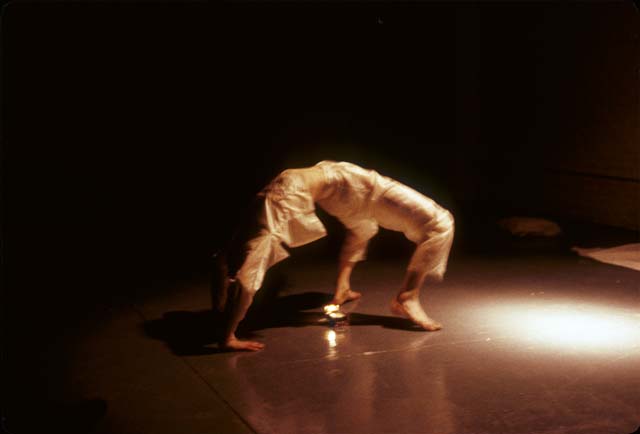 A color photo of a performance in which a person wearing cream colored pants and shirt is caught mid-back-bend.