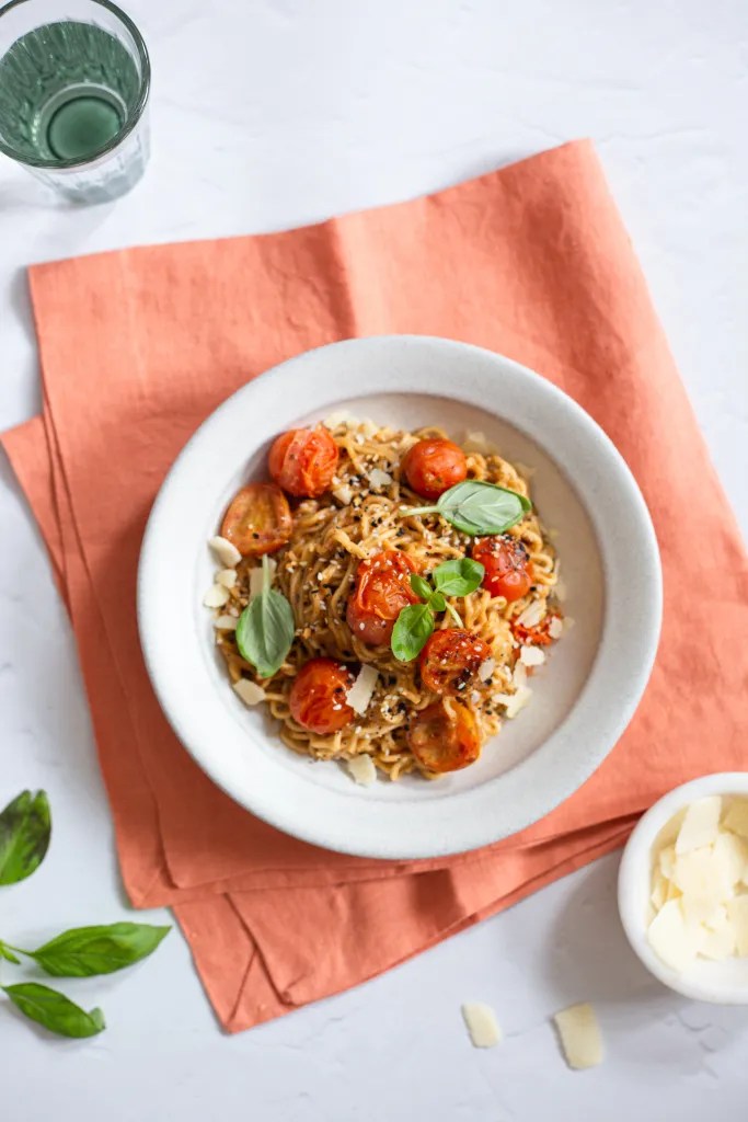 a bowl of burst tomato miso noodles garnished with shaved parmesan and fresh basil
