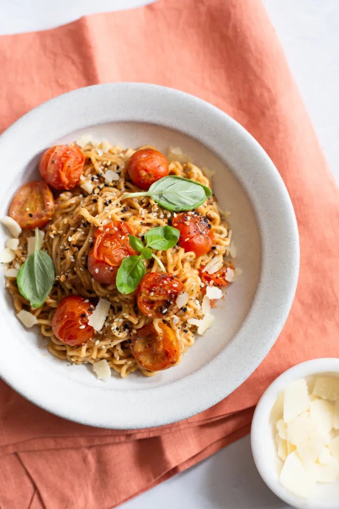 a bowl of burst tomato miso noodles garnished with shaved parmesan and fresh basil