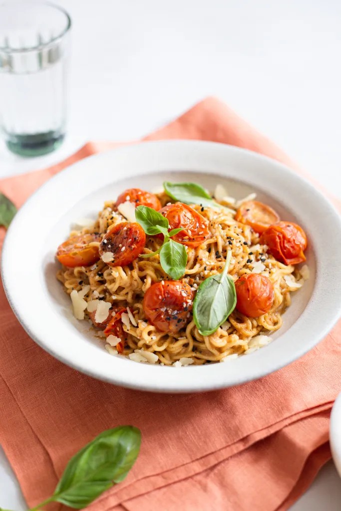 a bowl of burst tomato miso noodles garnished with shaved parmesan and fresh basil