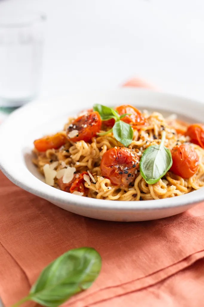 a bowl of burst tomato miso noodles garnished with shaved parmesan and fresh basil