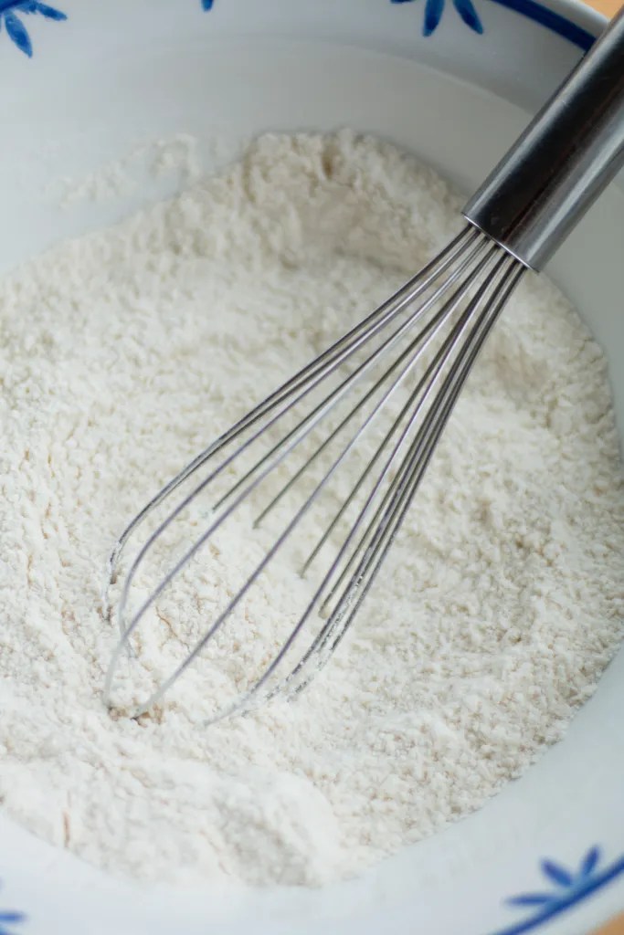 A metal whisk mixing flour, sugar, baking powder, baking soda, and salt in a clear glass bowl.