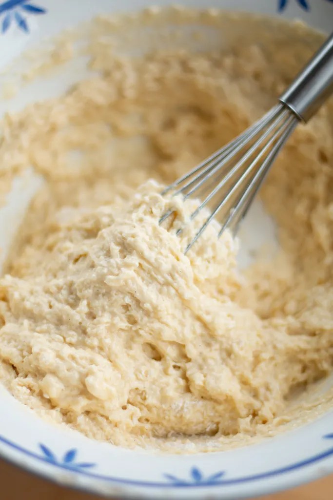 A whisk folding the wet buttermilk mixture into the dry flour ingredients to form a thick donut batter.