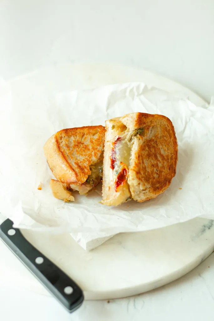 A sun-dried tomato grilled cheese sandwich cut in half, revealing melted cheese and capers.