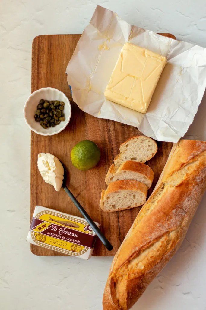 ingredients for easy sardine spread laid out on a cutting board, including butter, sardines, lemon, and capers.