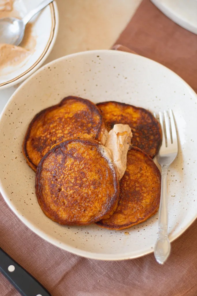 A tall stack of fluffy pumpkin pancakes on a white plate, topped with a melting pat of cinnamon brown butter.