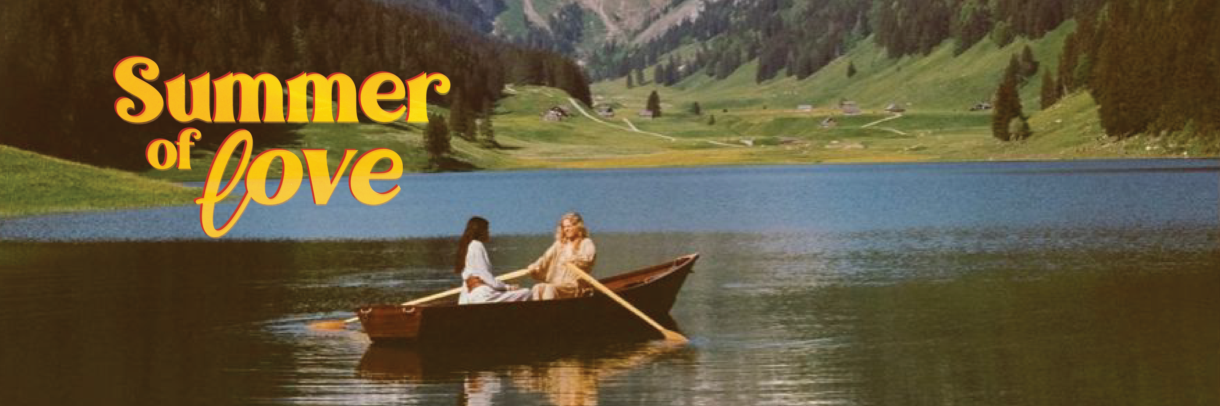 A panoramic banner image with a vintage, warm-toned aesthetic. In the center, two women in light-colored clothing sit in a small wooden rowboat on a calm, blue lake. The background features rolling green alpine hills, a distant forest, and mountains. In the upper left corner, the words "Summer of love" are written in a prominent, 1970s-style yellow script font with a red outline.