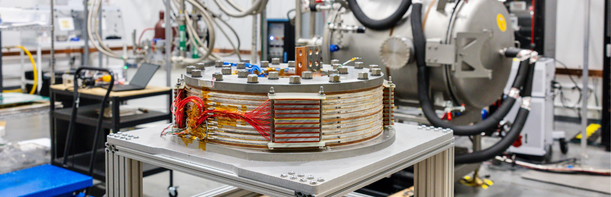 A full-scale planar shaping coil for Thea Energy's Eos stellarator sits on a laboratory bench, showing the circular winding geometry and instrumentation wiring used during magnet performance testing.