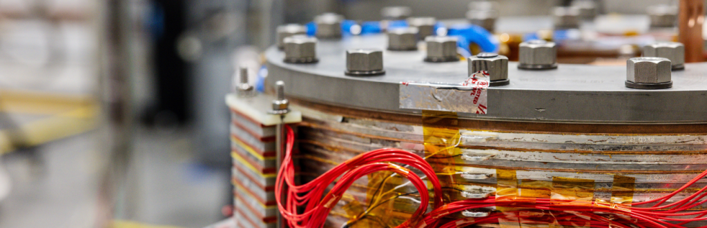 A modular high-temperature superconducting magnet coil, its copper and HTS tape windings visible in cross-section beneath a bolted end plate, representative of the hardware Thea Energy is scaling toward volume HTS magnet manufacturing.
