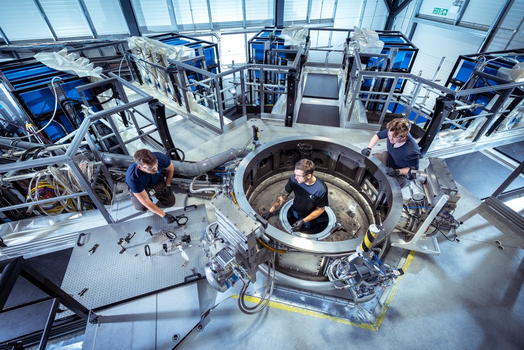 First Light Fusion engineers working inside the open target chamber of a pulsed power fusion facility, with capacitor banks and pulse-forming equipment visible in the background.
