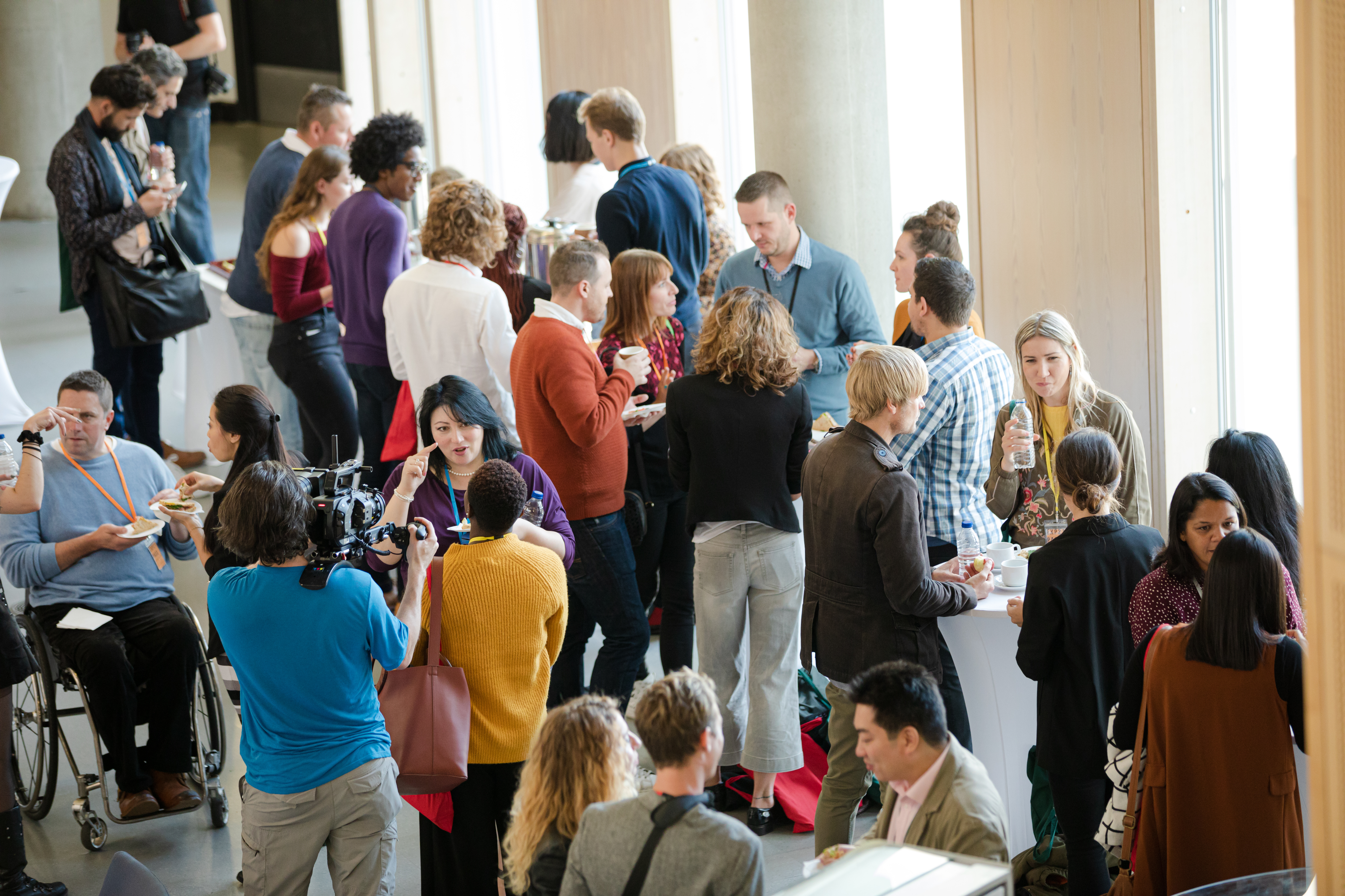 A gathering of 25+ adults standing around small banquet tables in a hall, socializing.