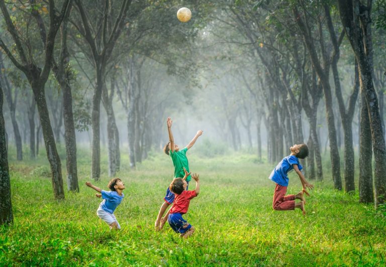 Four young boys play with a soccer ball in a grassy, tree-lined field.