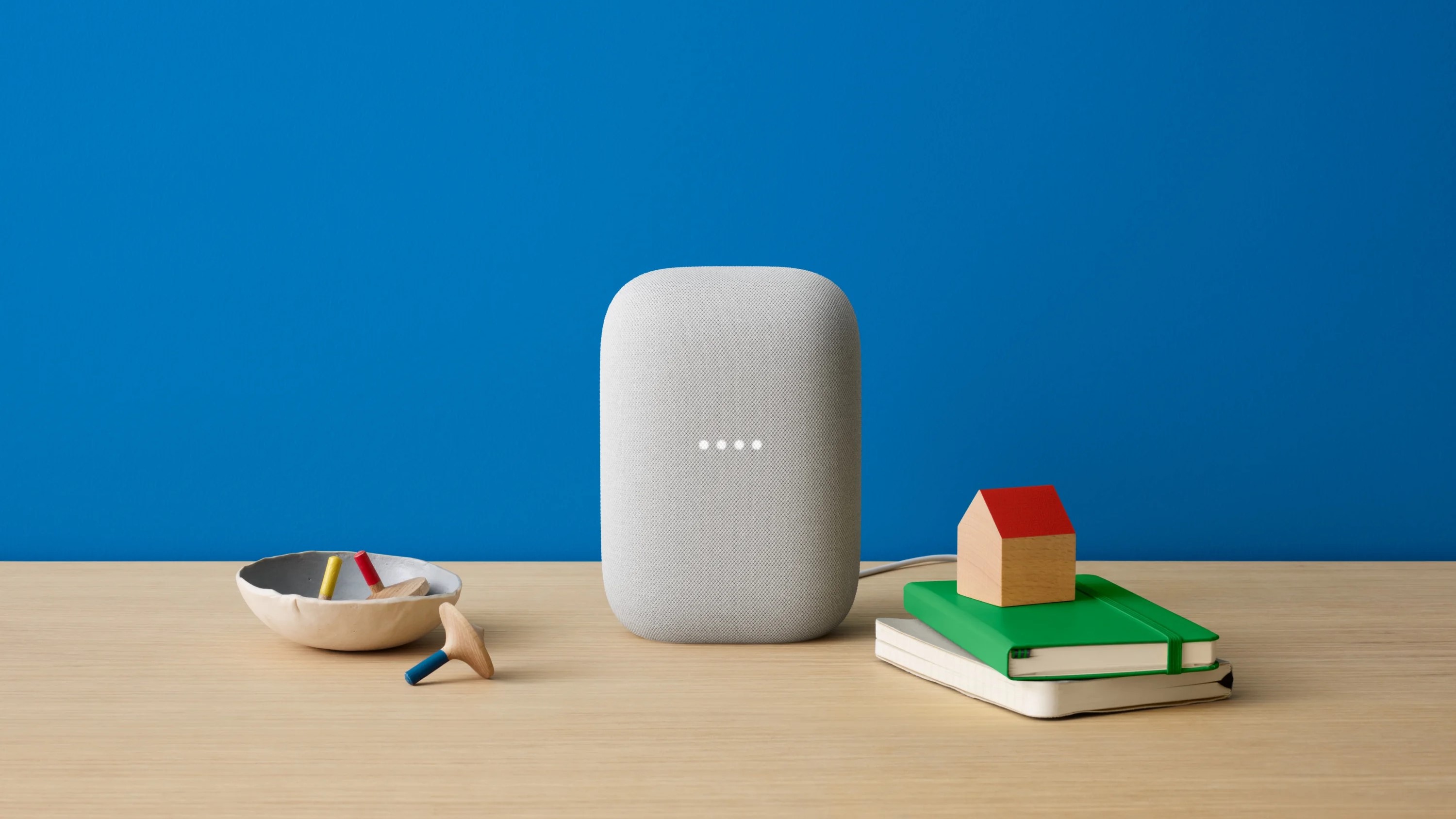 This image shows a smart speaker, likely a Google Nest Audio, positioned on a wooden surface with a minimalist setup. The presence of the bowl with wooden toys and the notebooks with a wooden house model suggests a blend of modern technology with simple, playful, and educational elements, making it visually appealing and relevant for showcasing smart home devices in a family-friendly environment.