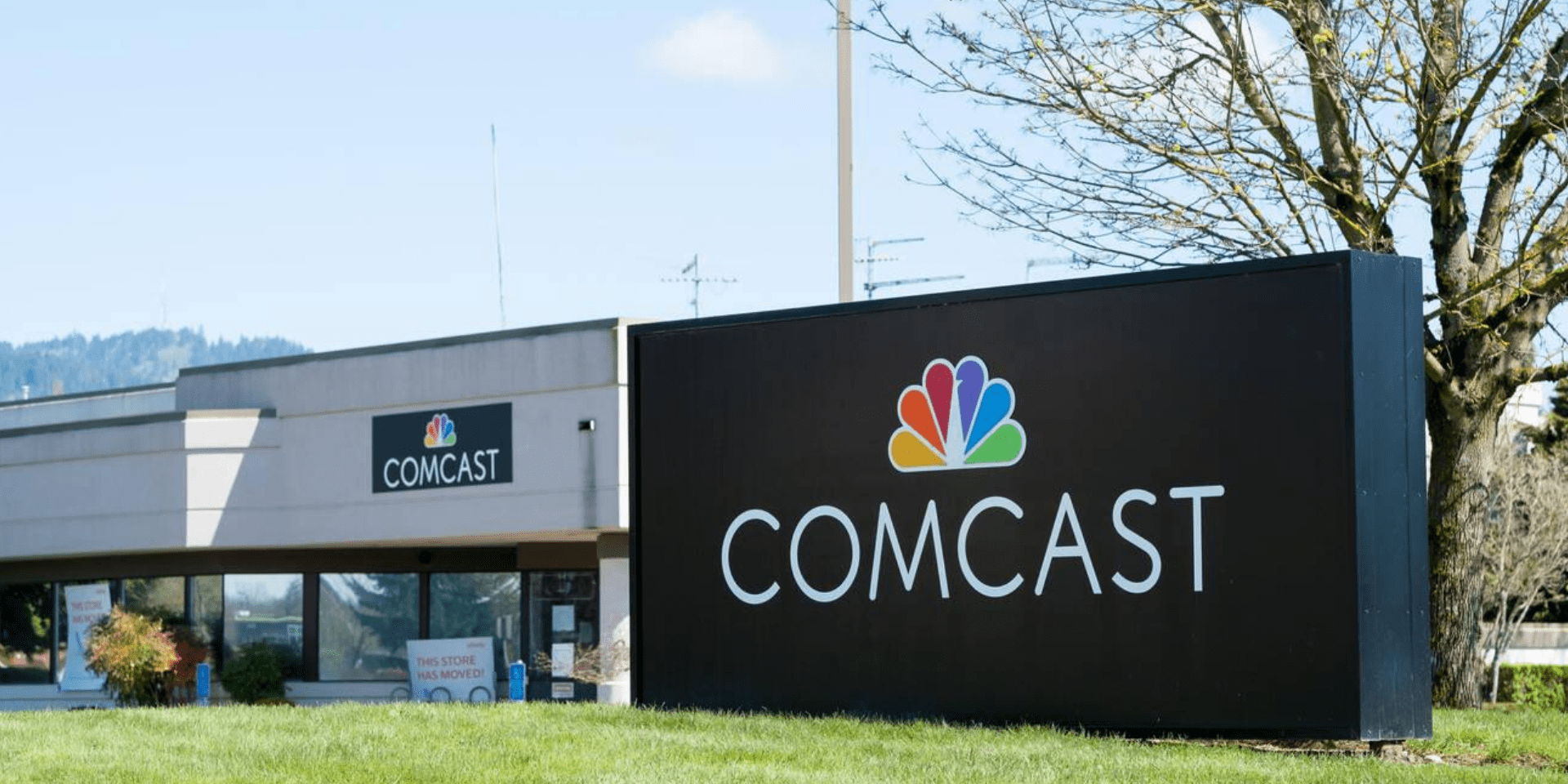 The image features a large, prominent sign with the word ‘COMCAST’ in capital letters, accompanied by the colorful NBC peacock logo to the left. The sign is situated outdoors in front of a building with a beige facade and several visible windows. The building also has a smaller sign displaying ‘COMCAST’ next to an entrance. In the background, there are trees and a clear blue sky, suggesting a sunny day without clouds.