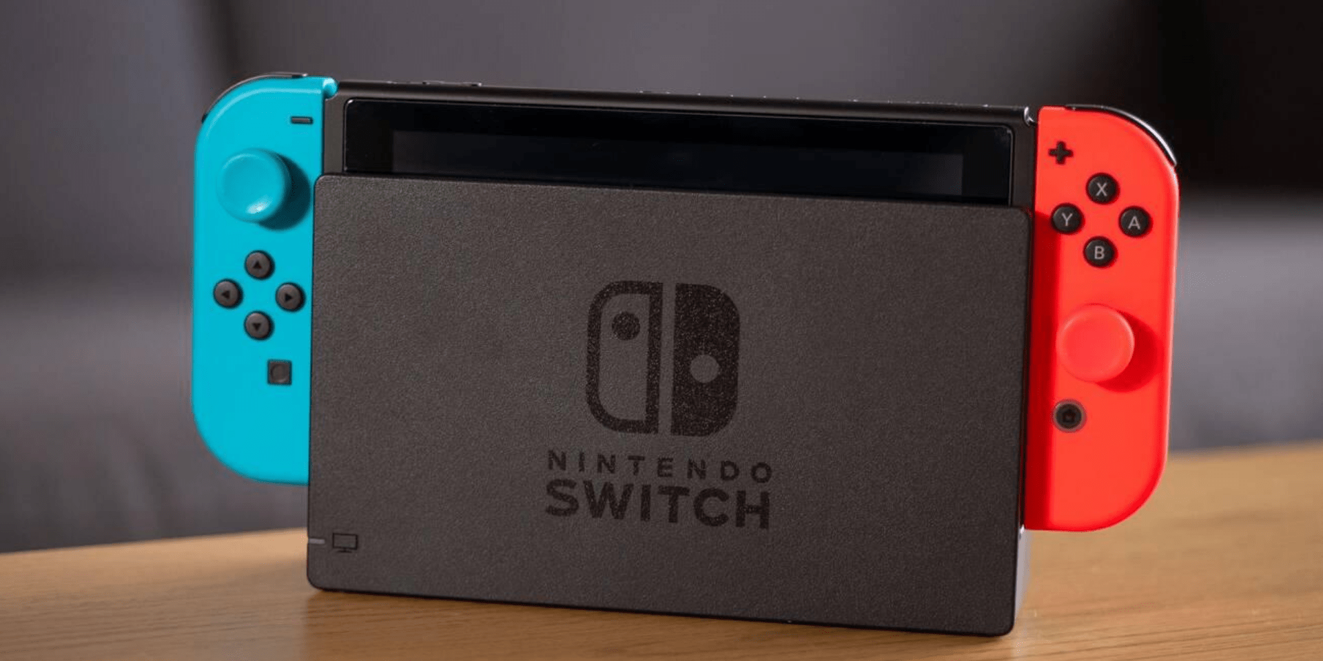 The image features a Nintendo Switch console in its dock. The console has a pair of Joy-Con controllers attached, one neon blue on the left and one neon red on the right. The dock is gray with the Nintendo Switch logo in white at the center. The background is blurred but appears to be a wooden surface with a soft-focus room setting.
