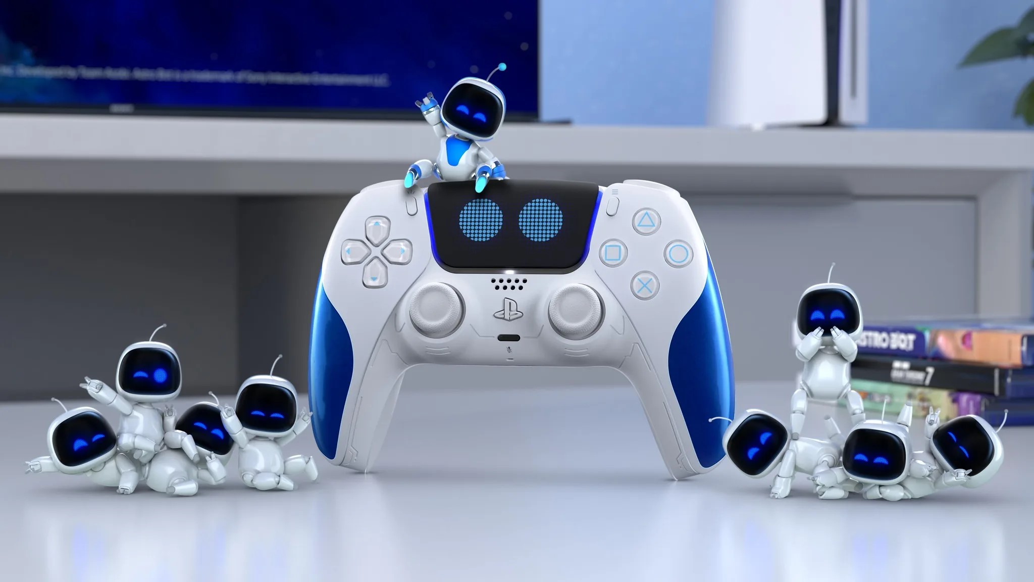 The image features a collection of small, white robot figures with blue accents, resembling the Astro Bot character. These figures are arranged around a white with black and blue accent PlayStation DualSense Wireless Controller Astro Bot Limited Edition that is centrally positioned. Behind the controller, there is a stack of books and a blurred monitor on a desk in an office setting. The robots appear to be interacting with each other and the controller, creating a playful scene that showcases PlayStation gaming accessories.