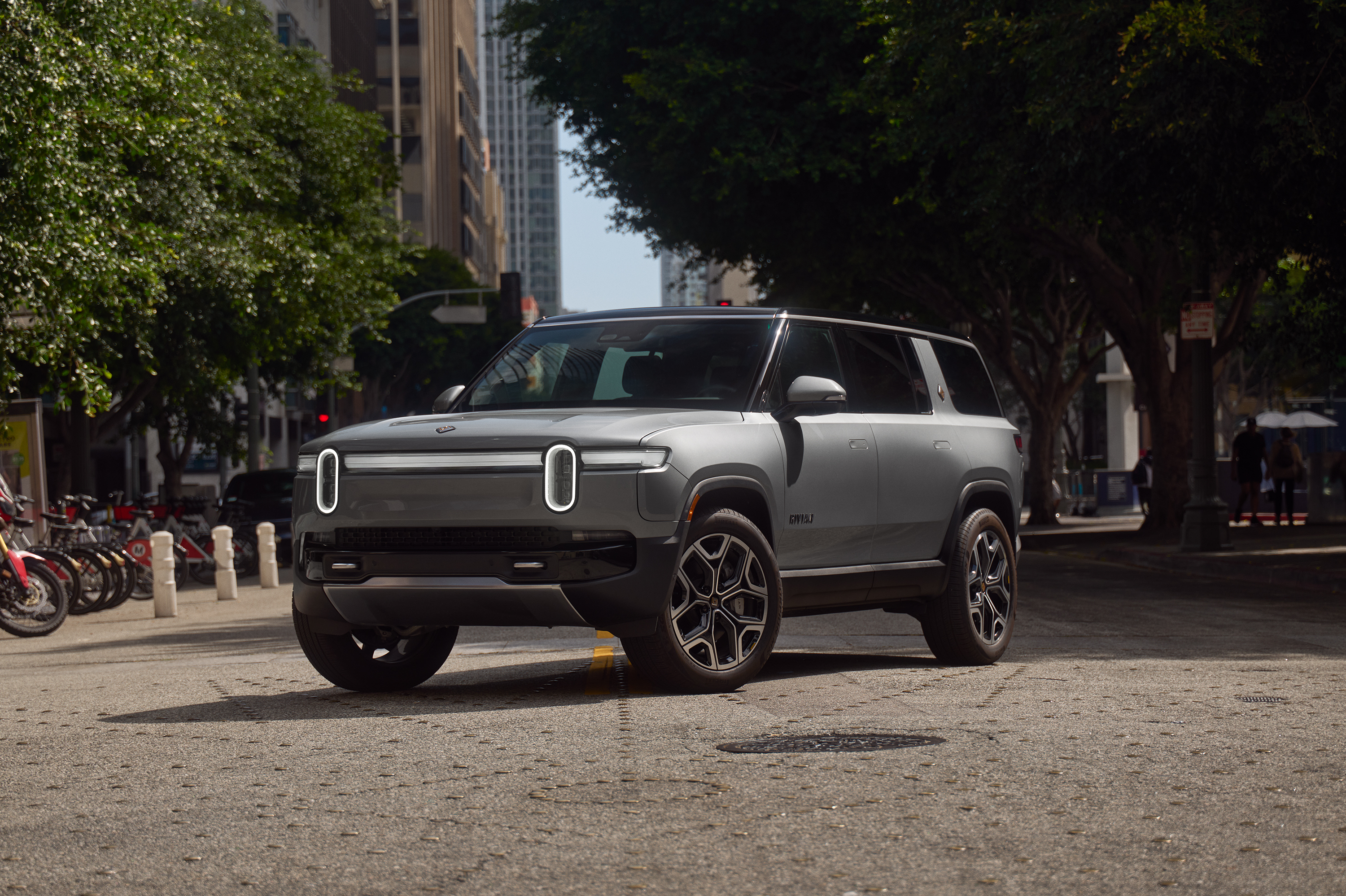 Silver 2027 Rivian R1S electric SUV parked on a city street with trees and buildings in the background.