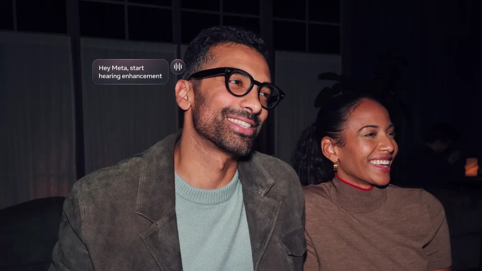 Smiling man wearing Meta AI glasses beside a smiling woman, with a prompt reading: "Hey Meta, start hearing enhancement".