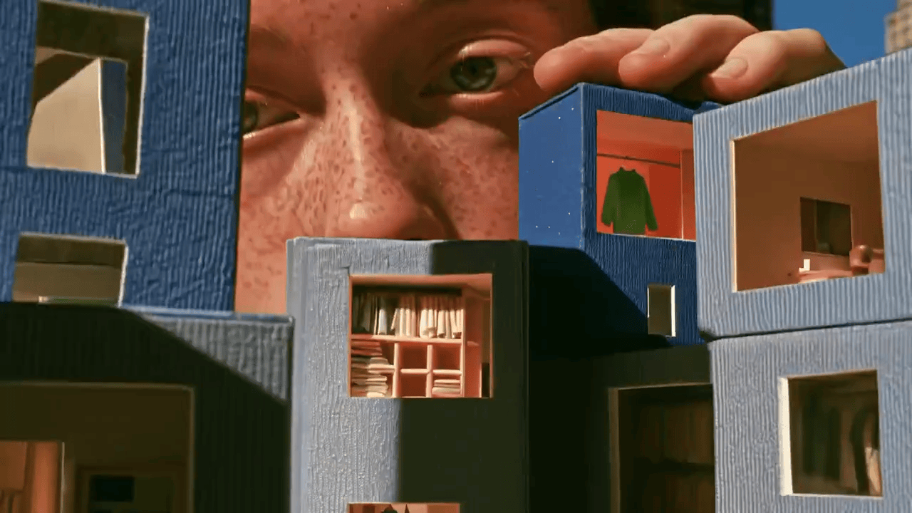 A close-up view of a person with freckled skin peering down into a miniature, dollhouse-style city made of blue modular cubic rooms. The tiny illuminated cut-out interiors showcase miniature domestic scenes, including a room with a green jacket hanging on a rack, a study lined with bookshelves, and small furnished living spaces.
