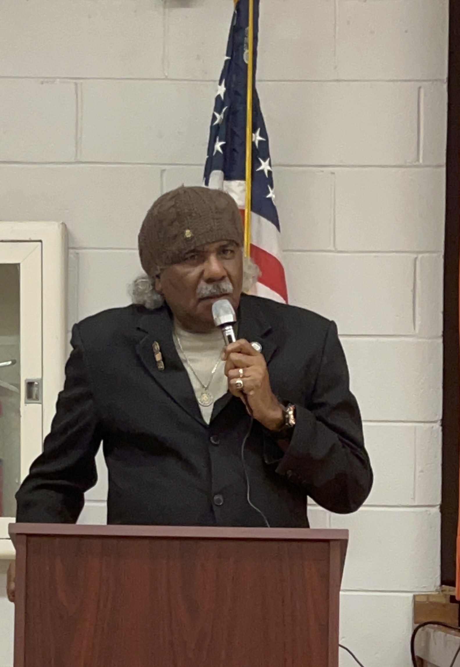 A man wearing a knit cap and dark jacket speaks into a microphone from behind a wooden podium, with an American flag behind him.