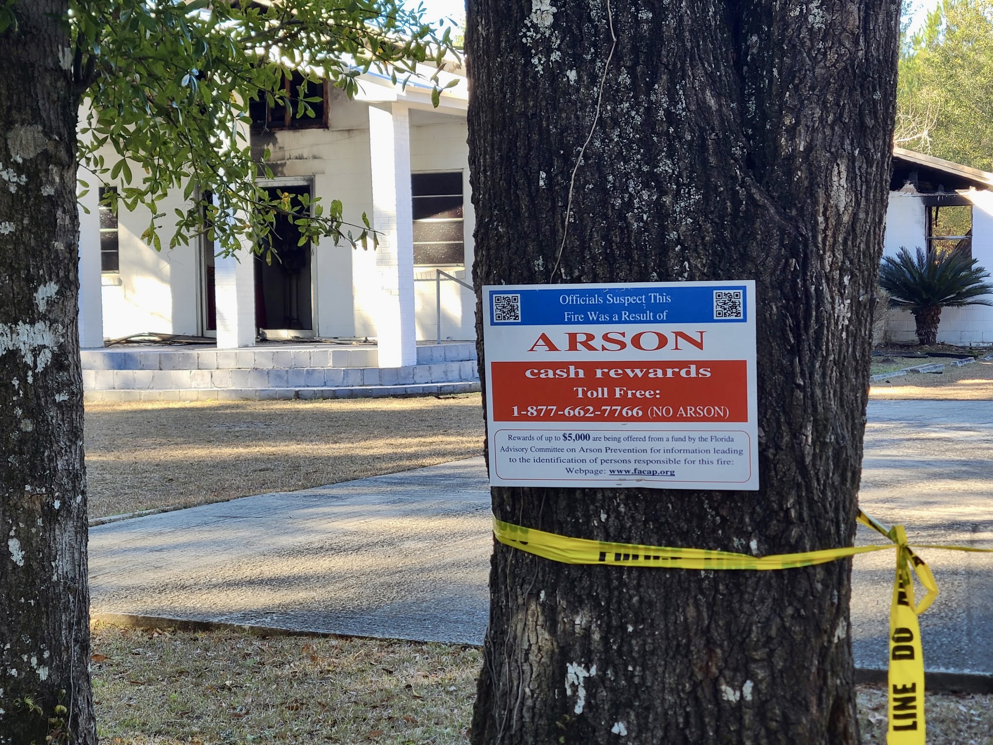 Arson investigation sign attached to a tree with yellow caution tape, positioned in front of a fire-damaged church building in the background.