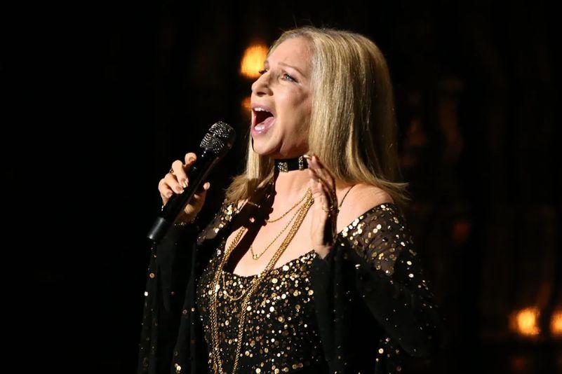 Barbra Streisand cantando hoje em dia, com cabelos loiros e roupas brilhantes, em apresentação.