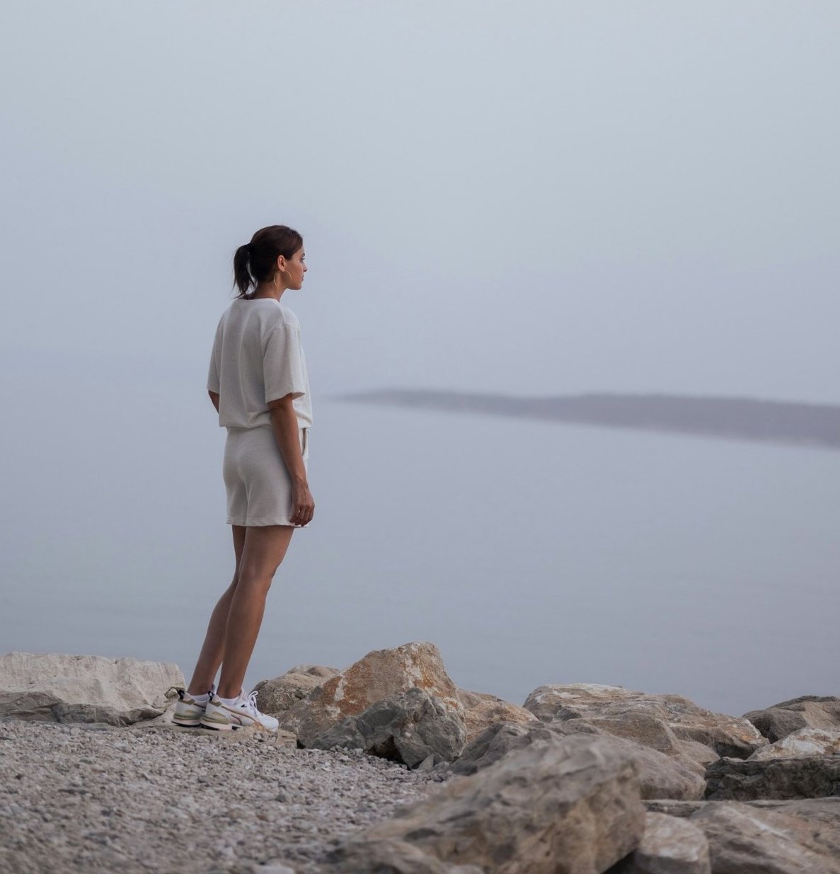 Person standing on rocky shoreline looking out over calm water, symbolizing reflection and navigating life transitions with Gentle Reframe.