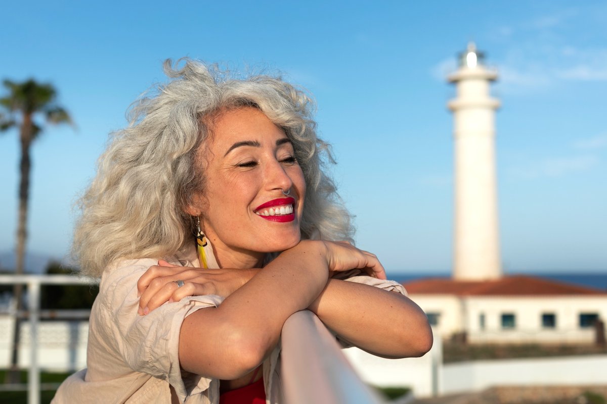 miling woman with silver hair leaning on a railing near a lighthouse, symbolizing clarity, guidance, and empowerment through Gentle Reframe MAP sessions.