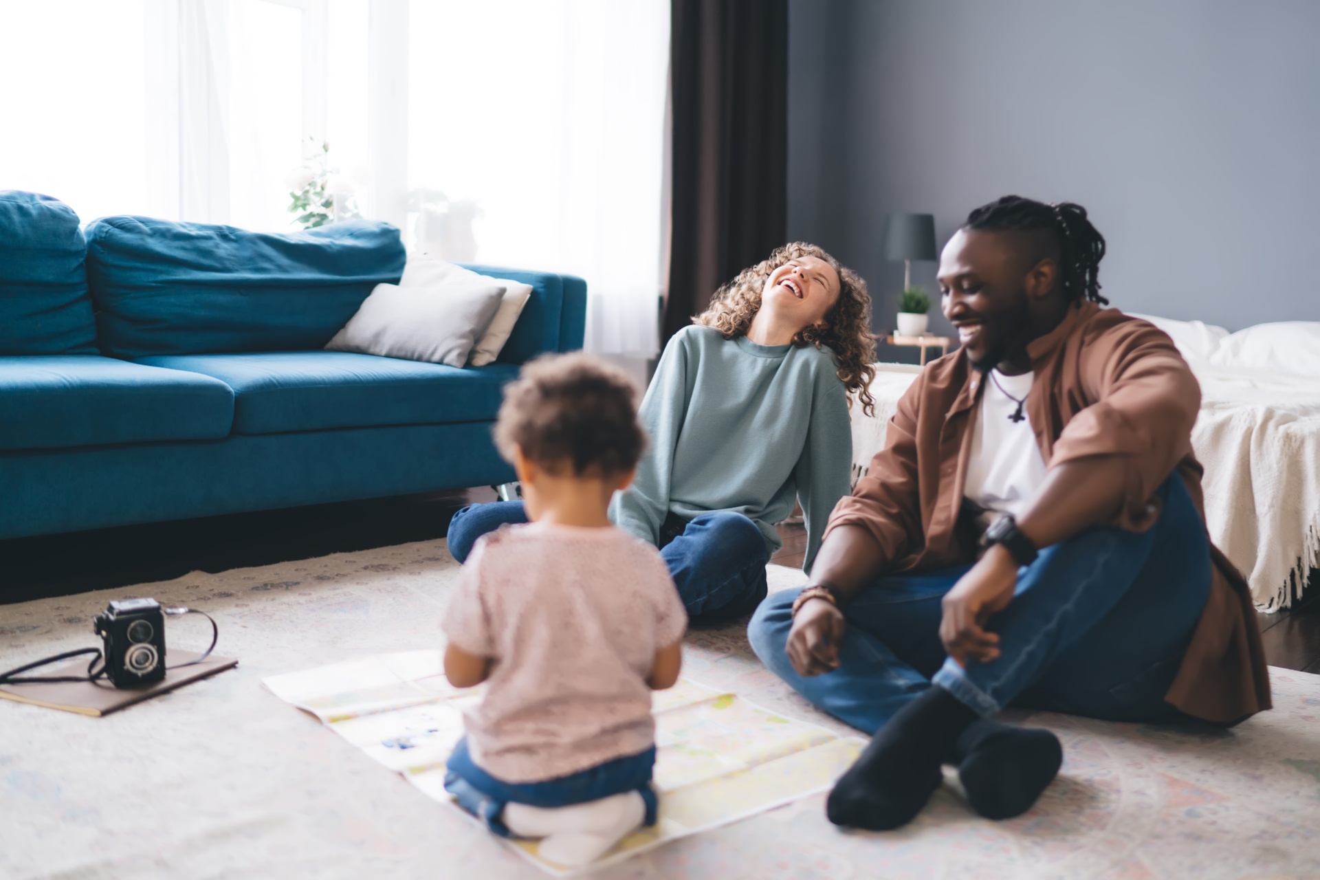 A pair of parents sit and play with their young child on a rug on the floor next to a blue sofa while smiling.