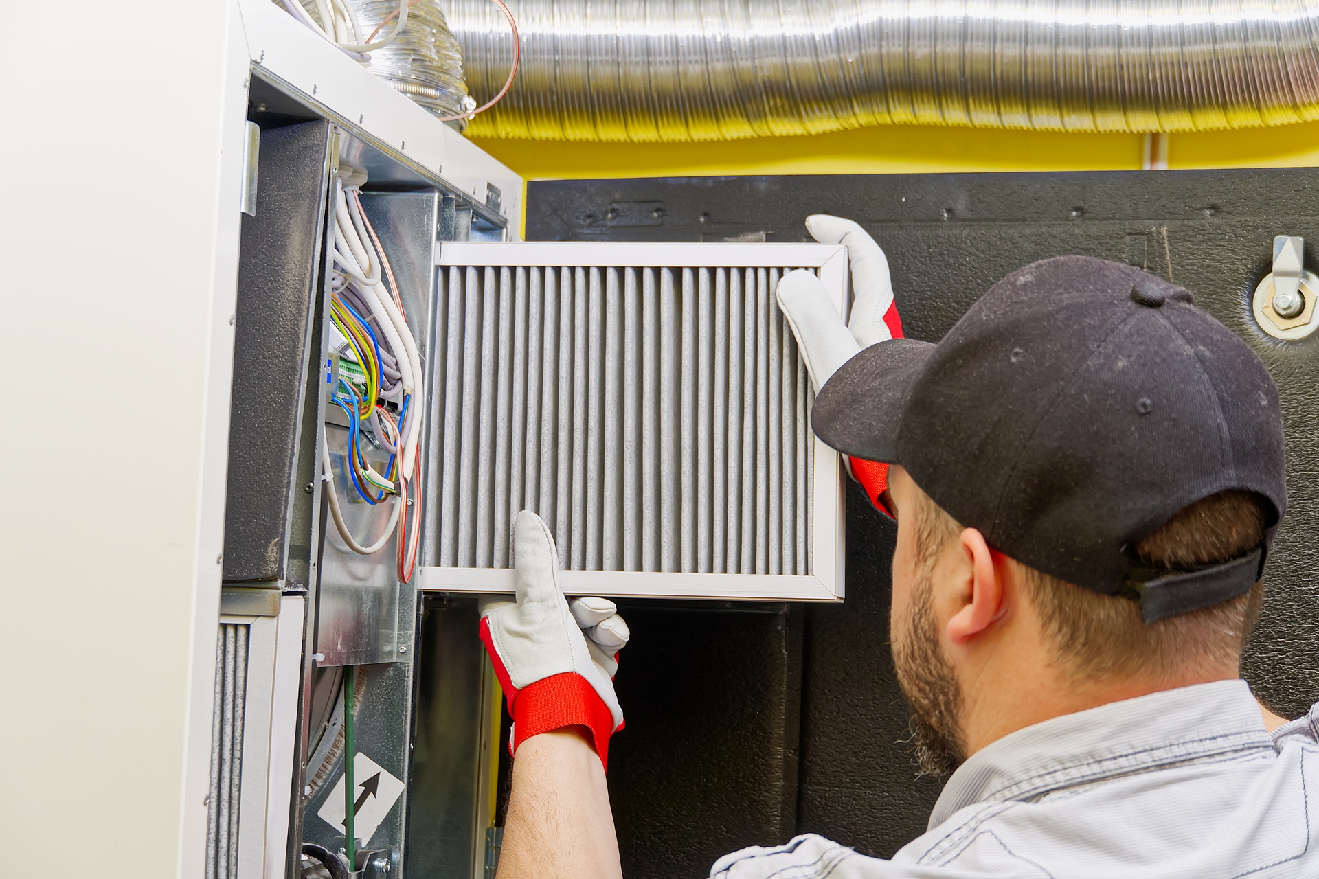 A man in a black hat, gray shirt, and white and red gloves installs a clean air filter in a central ventilation system.