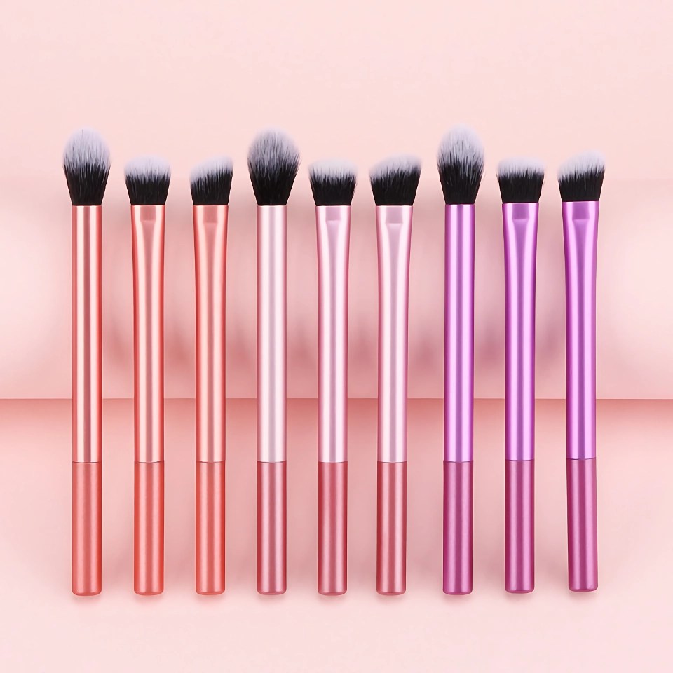 Row of eight metallic makeup brushes with pink-to-purple handles and fluffy white bristles against a pink background, reflected on the surface.