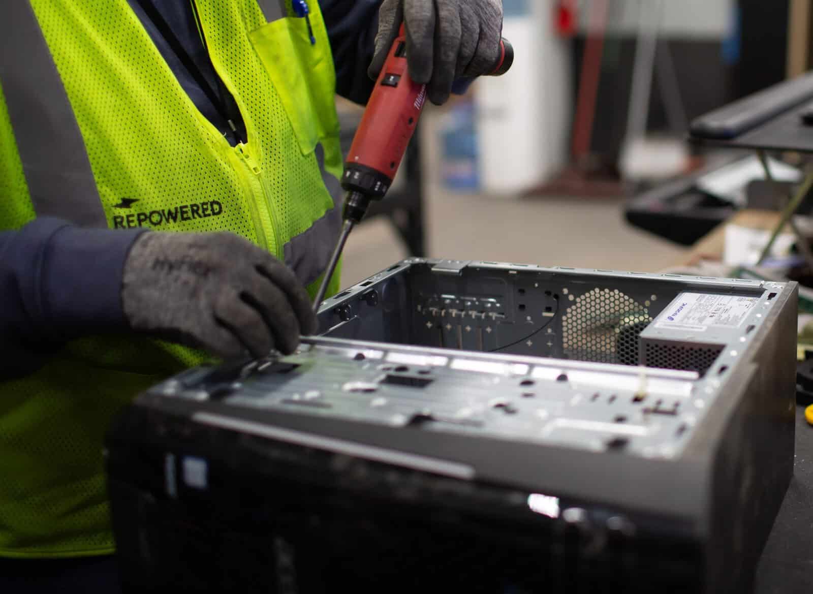 Technician in a high-visibility vest repairing a computer with a screwdriver in a workshop setting.