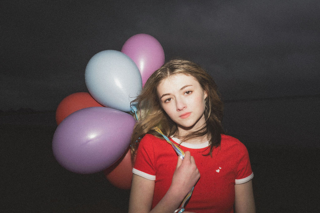teenage girl in red shirt holding 4 pastel balloons in front of dark sky