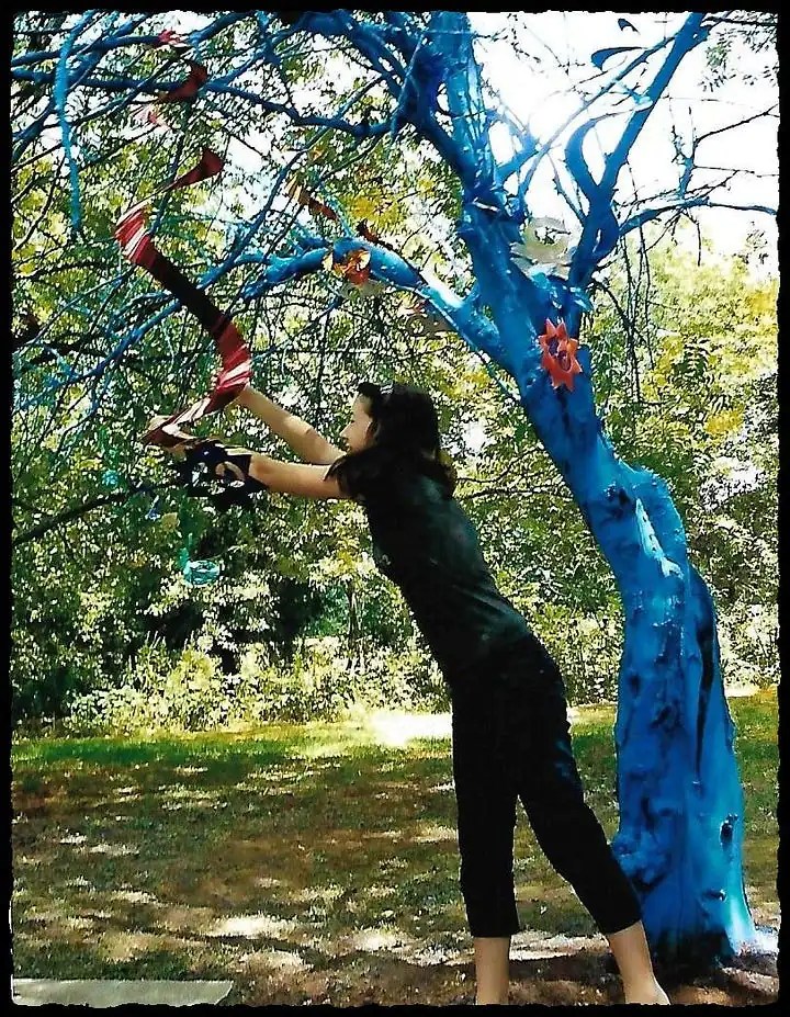 A young woman adds decoration to a dead apple tree that has been painted bright blue.