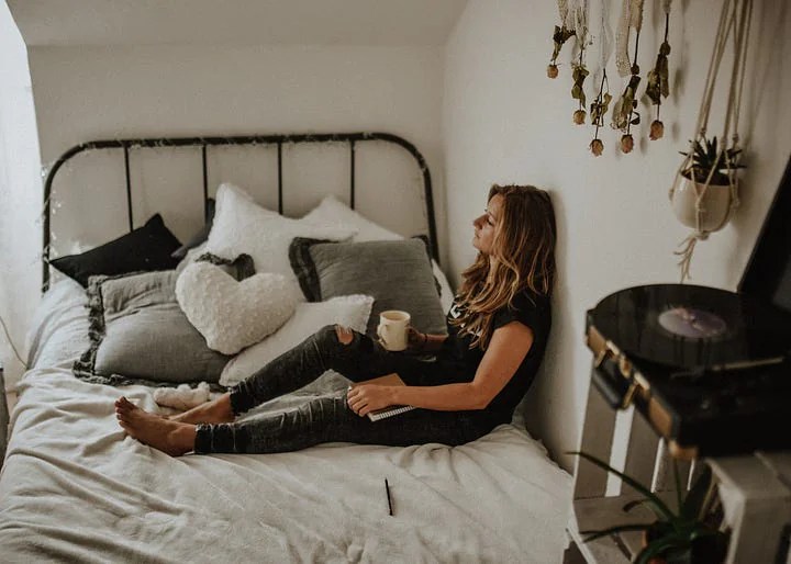 A young woman rest on an antique iron bed sideways with her back against the wall. There is a record playing, and plants showing. She has a journal and a cup of coffee or tea. She seems sad, maybe she is grieiving the loss of a baby.