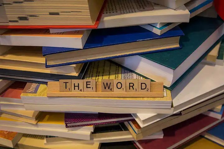 On an uneven stack of colorful hardback books rest scrabble letters that spells out The Word.