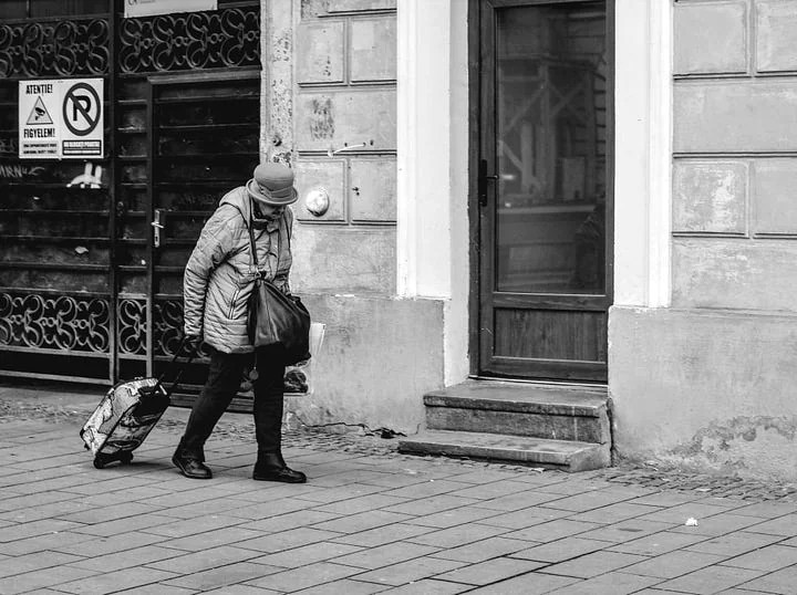 An elder woman struggles to pull her suitcase down the street, prehaps she is looking for shelter.