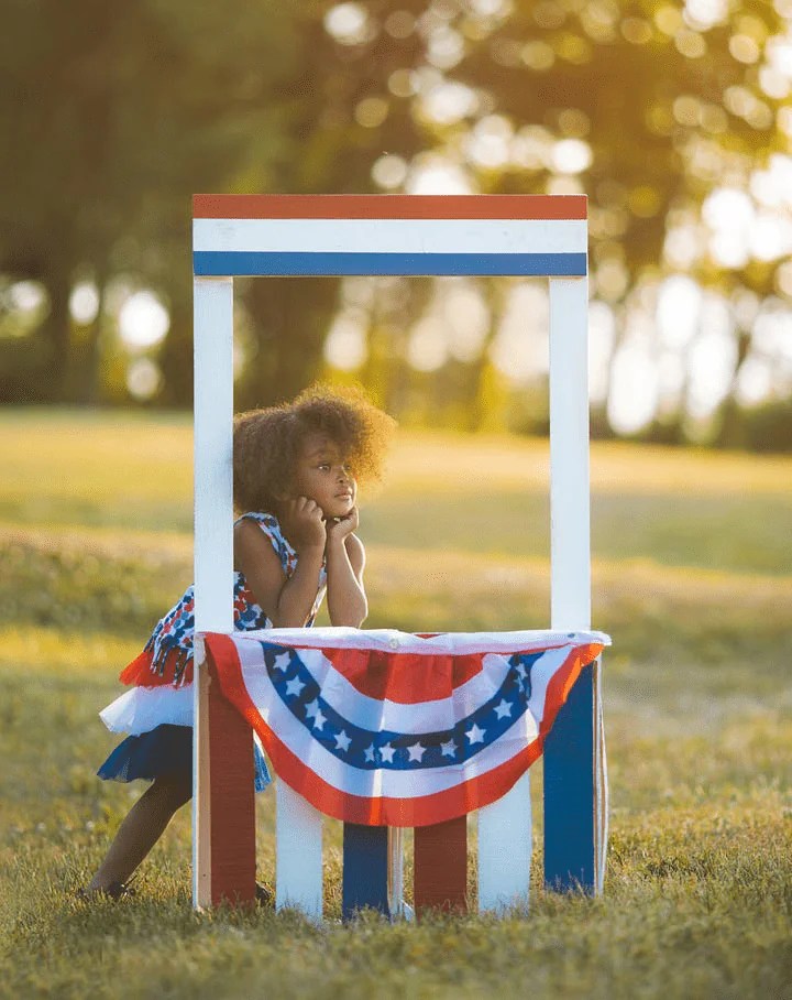 A small girl props up behind a red, whitle, and blue lemonade stand.
