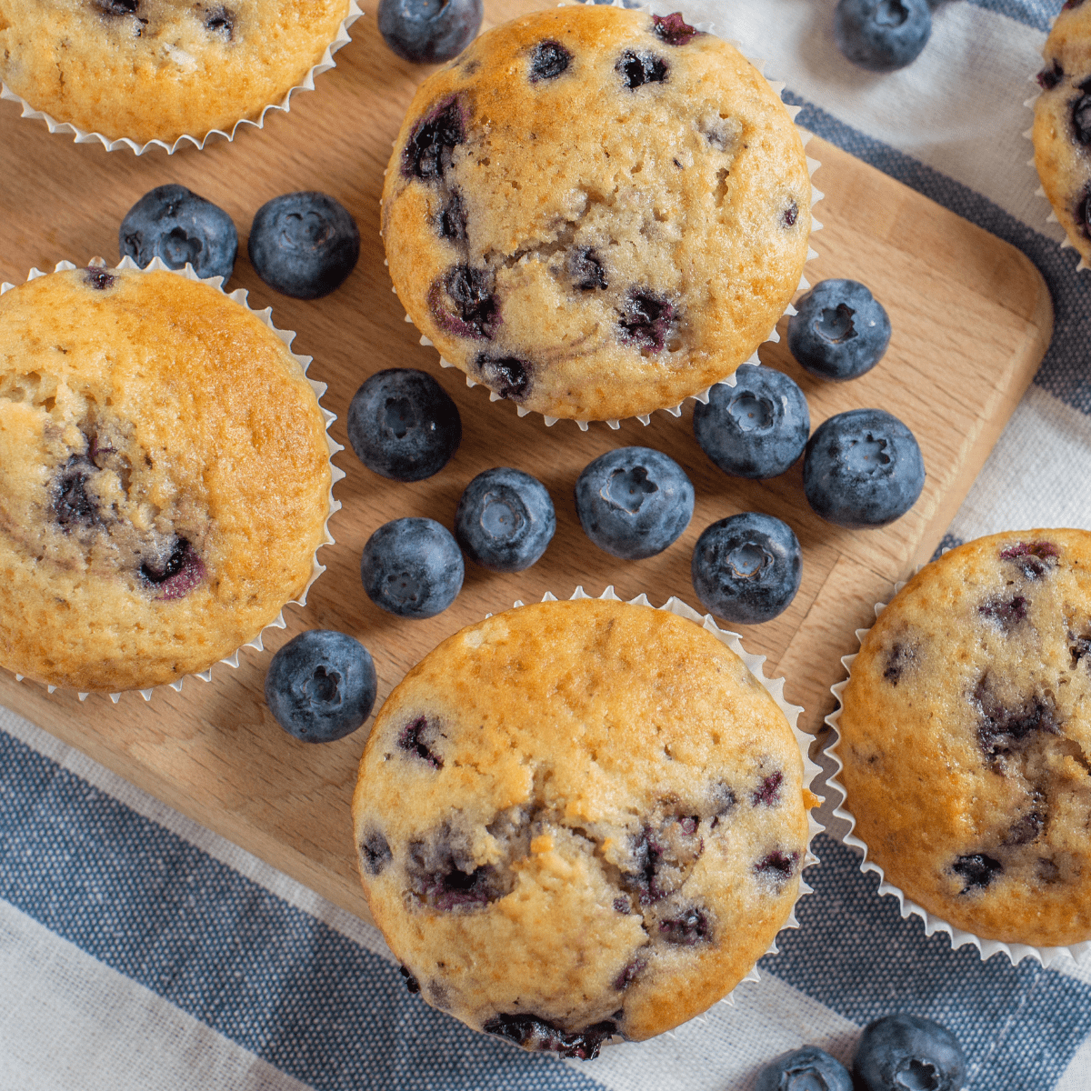 Five Blueberry muffins on a wooden cutting board with fresh blueberries scattered around.