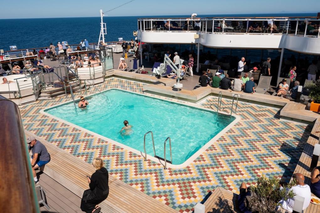 Sunny day at the Sea View Pool on Holland America’s ms Koningsdam, with passengers relaxing on loungers and enjoying the colorful tiled deck while sailing through the Alaska Inside Passage.