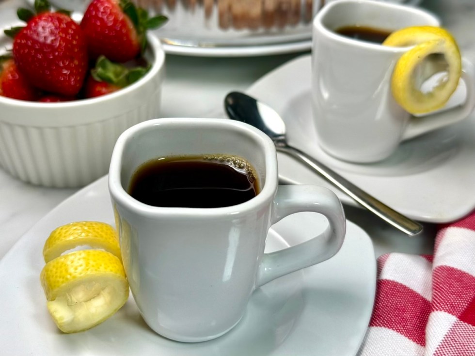 espresso in demitasse cups with lemon twists and strawberries and pastries in the background