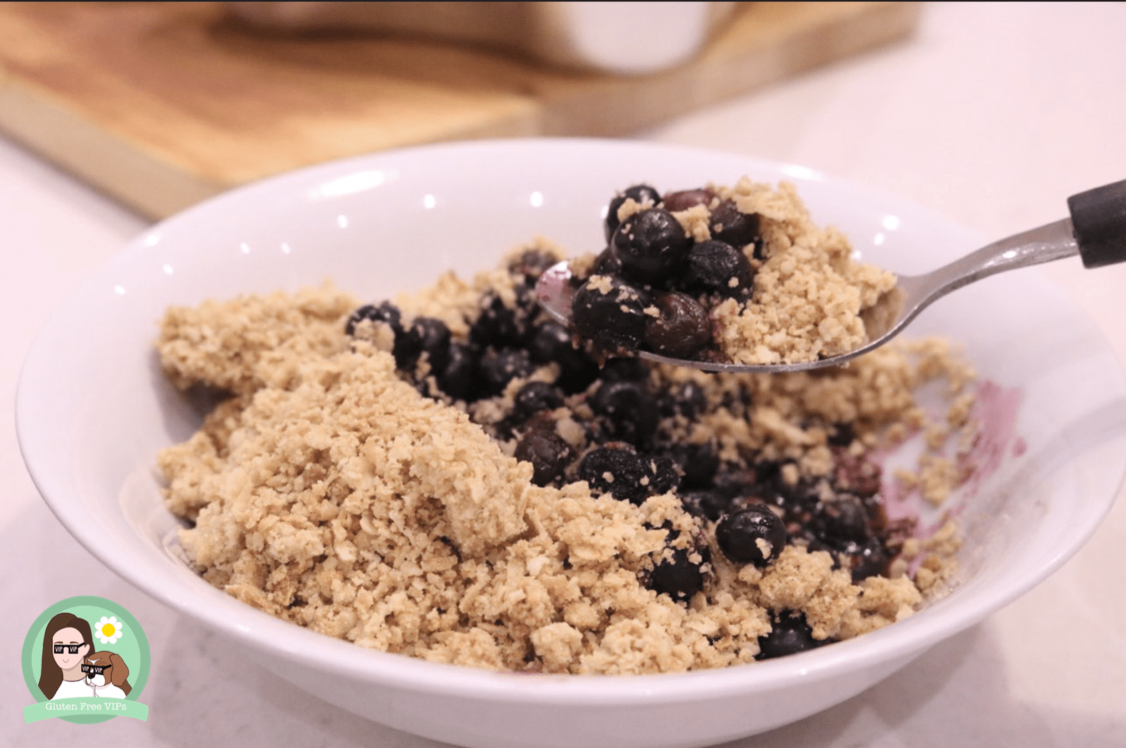 A golden, gluten‑free blueberry crumble in a baking dish, with bubbling blueberries beneath a crisp oat topping and a spoon ready to serve.