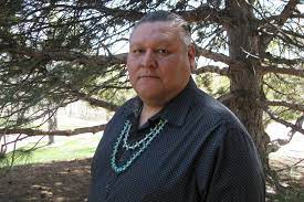 native american male wearing a blue shirt and a turquise necklace with a tree behind him