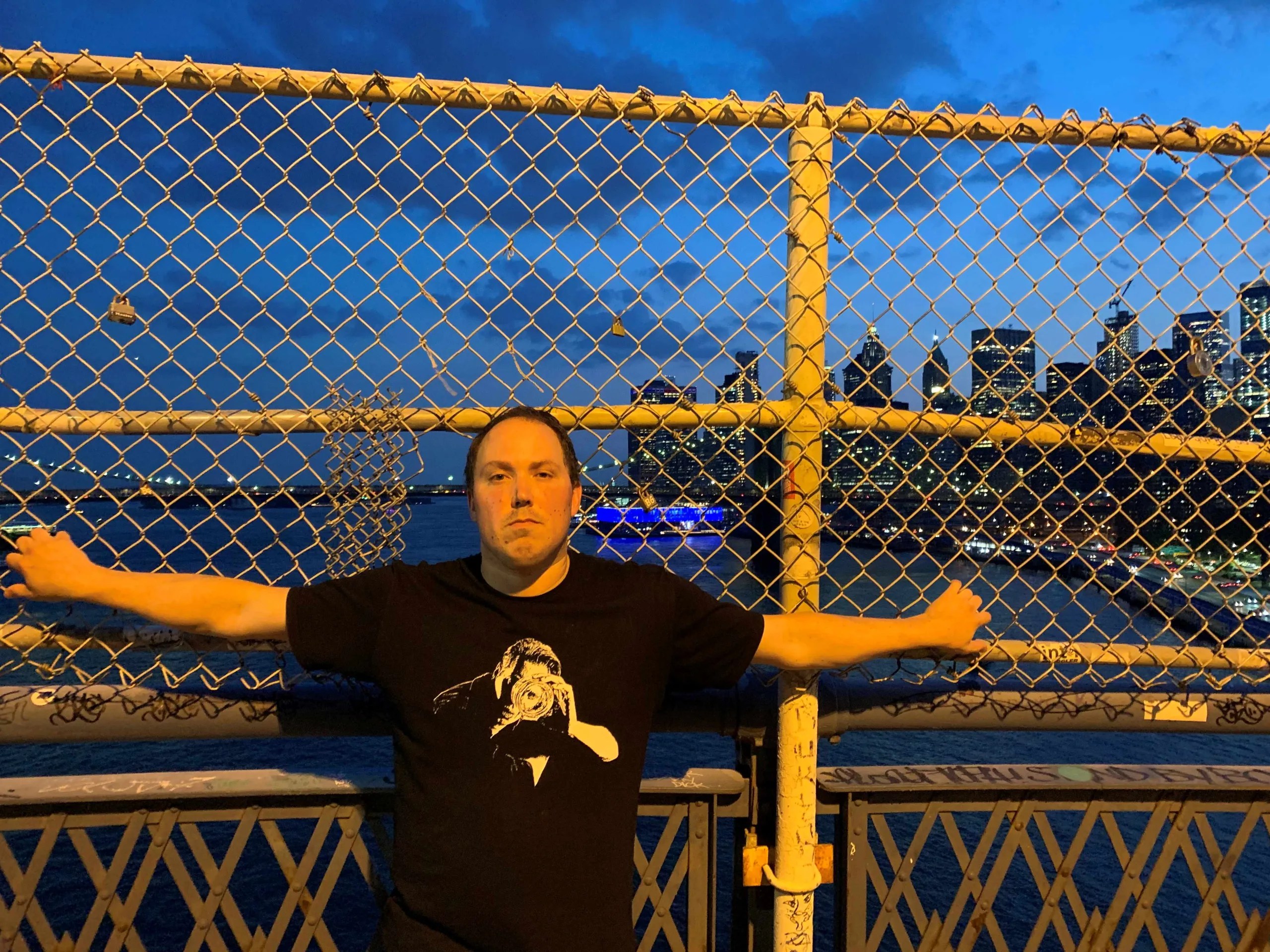 Man in a black t shirt with an image on it with his arms oustretched in front of a chain link fence