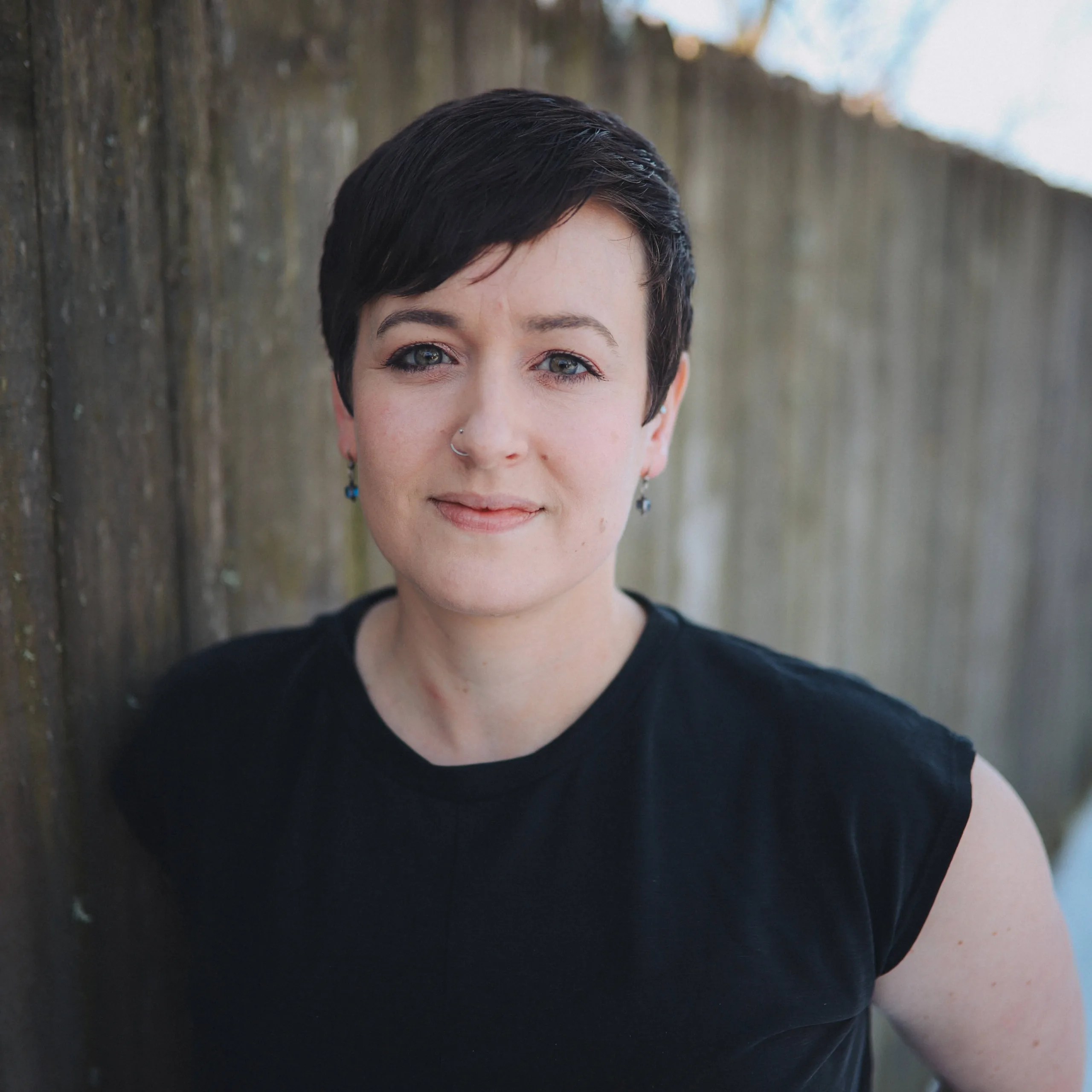 woman with short dark hair and black t shirt standing in front of a fence