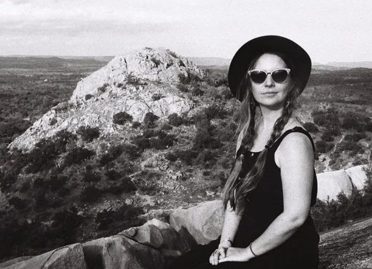 black and white photo of a woman with pigtails wearing a hat and sunglasses sitting in front of a mountain range